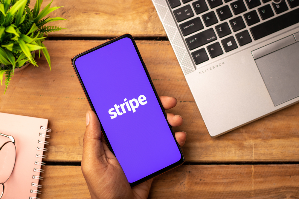 A hand holding a mobile phone with the purple Stripe logo on it next to a laptop computer and paper notebook.