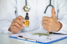 Doctor holding a dropper vial above a file with a hemp leaf and giving the thumbs up.