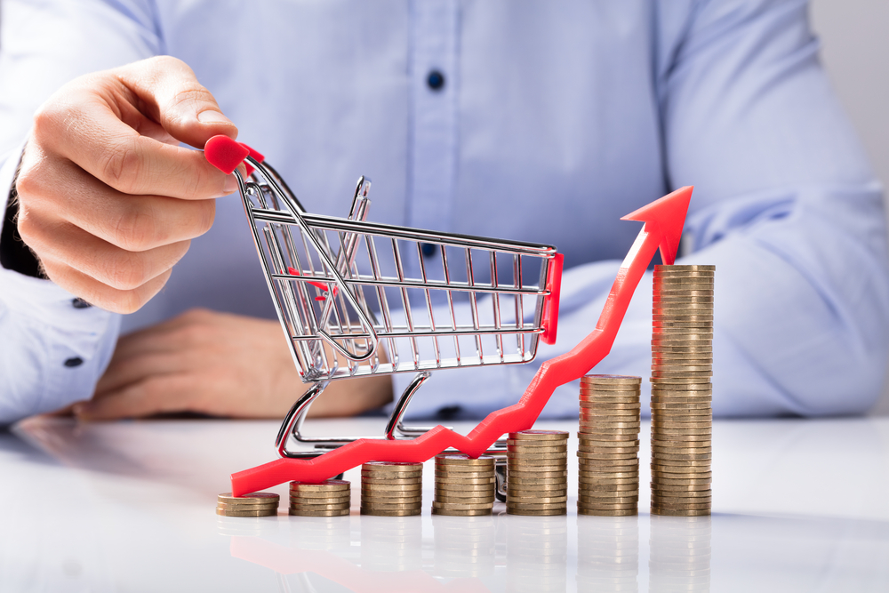 Hand pushing shopping cart on growing stacks of coins with red arrow pointing up.