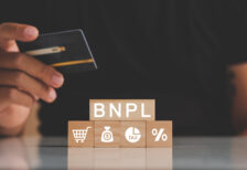 Man holding credit card above wooden blocks with BNPL, a shopping cart, money bag, tax symbol, and percent sign.