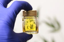 Hand wearing a glove holding a small glass container of oil reflecting cannabis leaves.