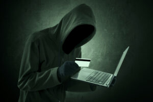Hooded man holding credit card in one hand and a laptop computer in the other.