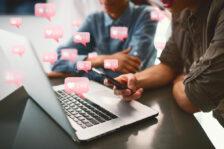 Two people in front of a laptop computer with social media "like", "follower", and "comment" icons floating up