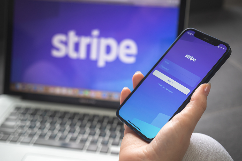 Hand holding a mobile phone showing the Stripe login page in front of a laptop screen showing a big Stripe logo.