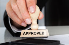 A hand holding a stamp that says "APPROVED" over a vat of ink.