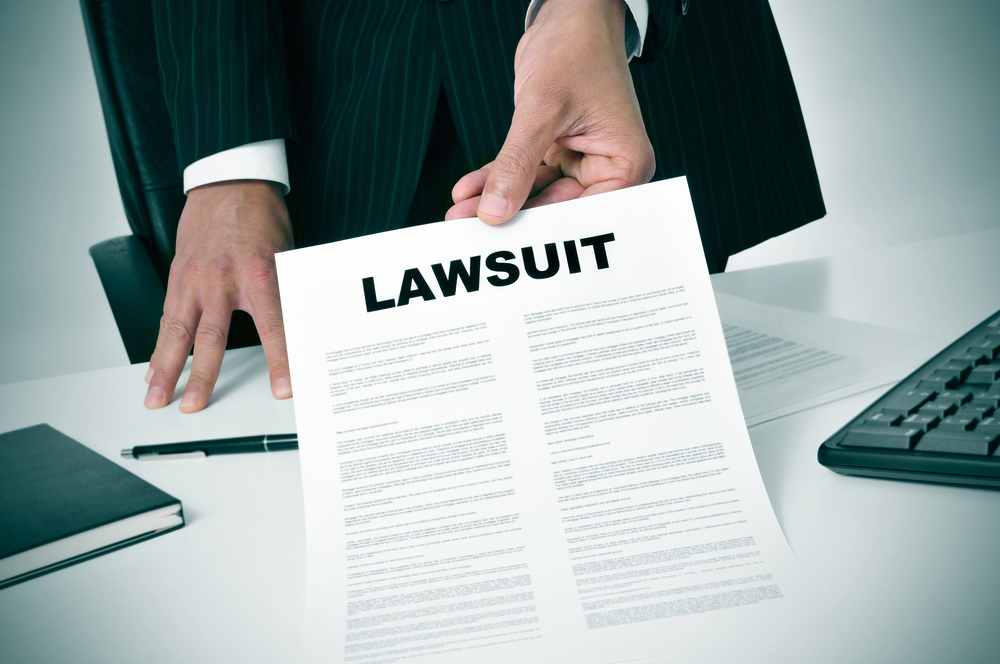 man wearing a suit passing a document with a bold "LAWSUIT" title across a desk