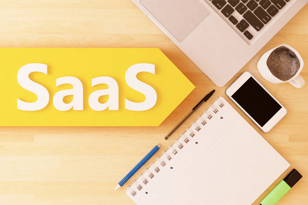 An arrangement of a laptop computer, a notebook, two pens, and an iPhone on the right with a big yellow arrow on the left that says "SaaS"