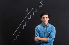 Man wearing a denim shirt standing in front of a blackboard that has a white chalk illustration of steps leading to a $. An arrow with a stick figure man points up the steps.