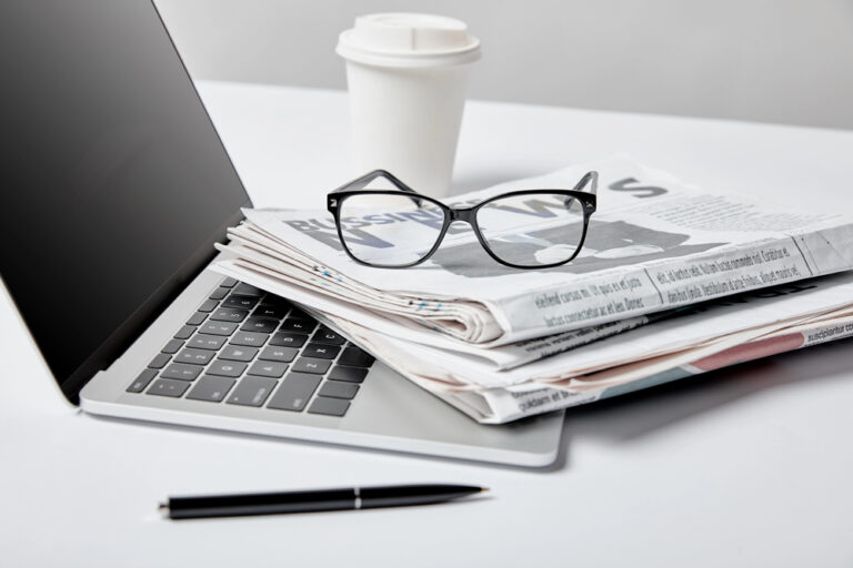 Selective focus of laptop with blank screen near business newspapers, glasses, pen and paper cup on white"